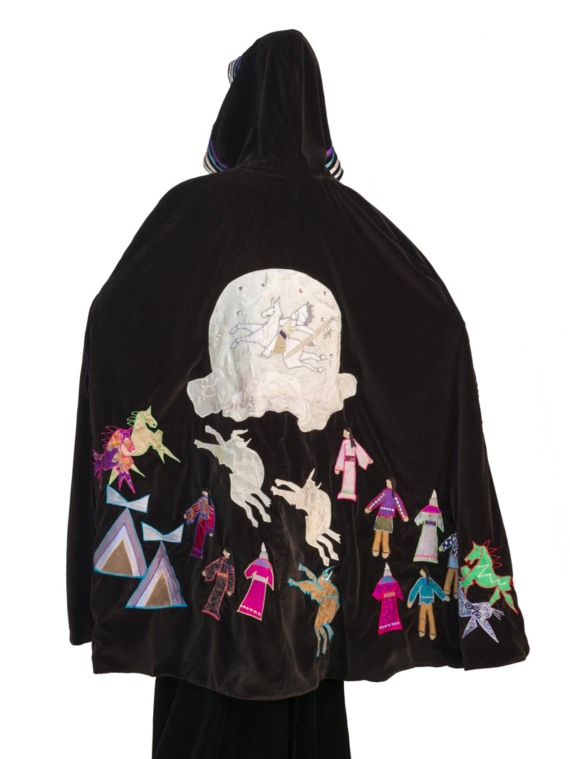 The back of a woman wearing a cloak with colorful designs, people and horses on it.