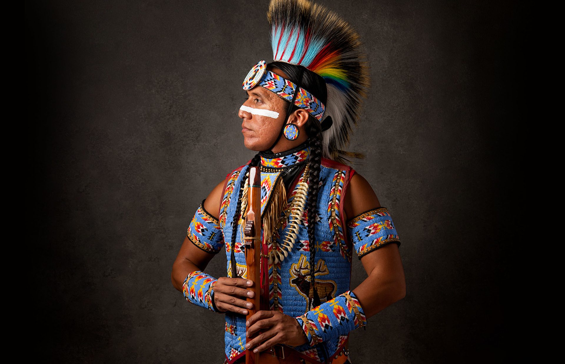A Native American man in a headdress and holding an instrument posing for a photograph.