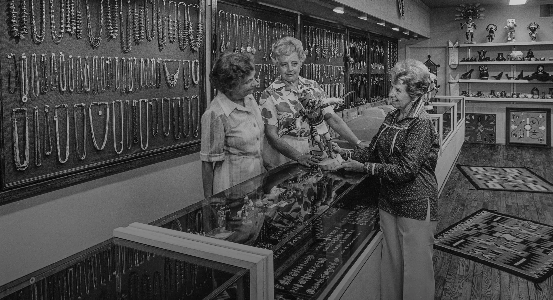 A black and white photo of three women talking at a jewelry case in a store where the walls are lined with necklaces and figurines.