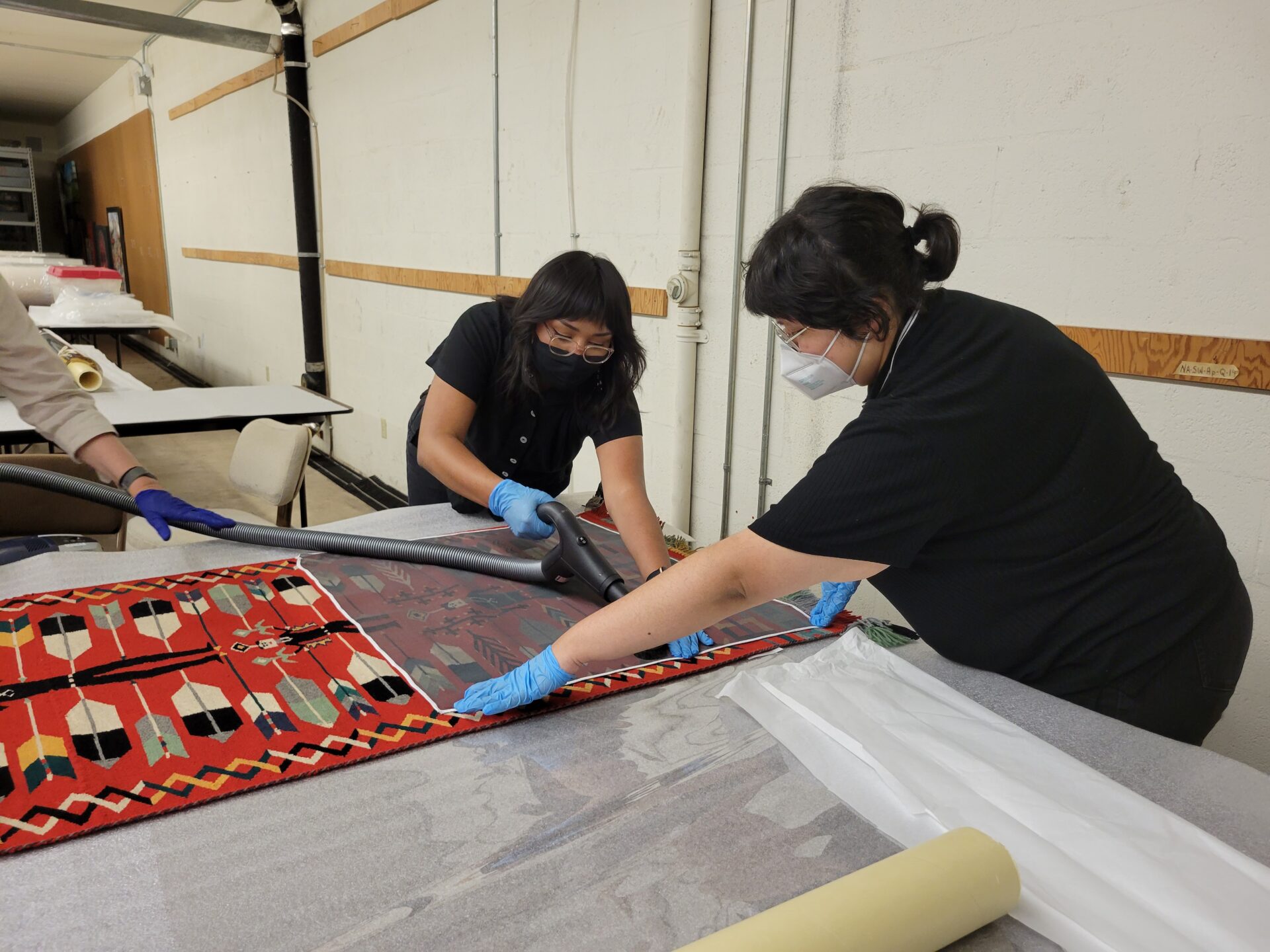 Two women wearing masks and gloves are cleaning a pictorial textile.