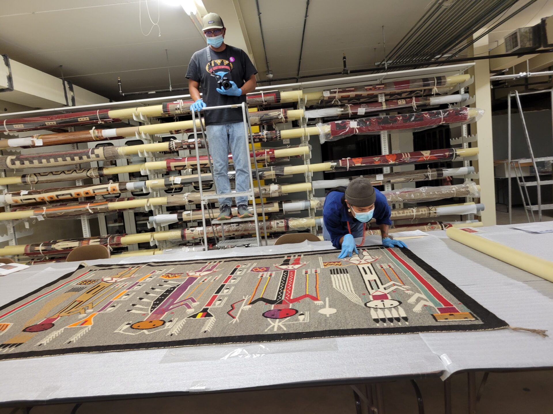 Two men inspecting a pictorial textile in a storage room.