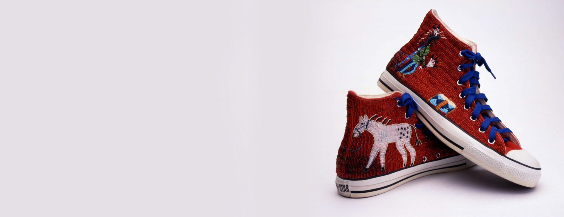 A pair of red, beaded Converse shoes with a white horse on them and blue shoelaces.