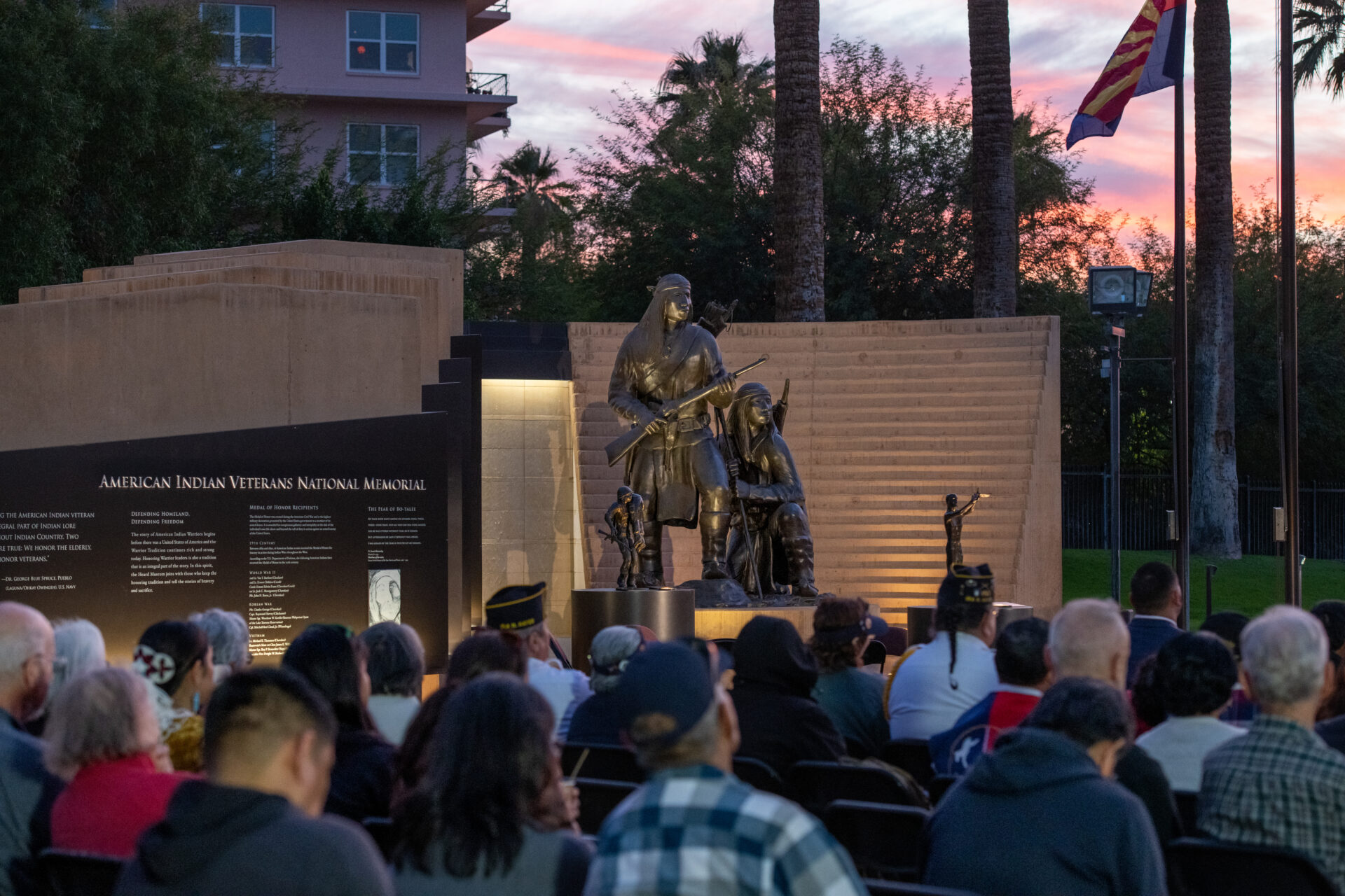 A sculpture of the American Indians Veterans National Memorial at dusk during a crowded event.