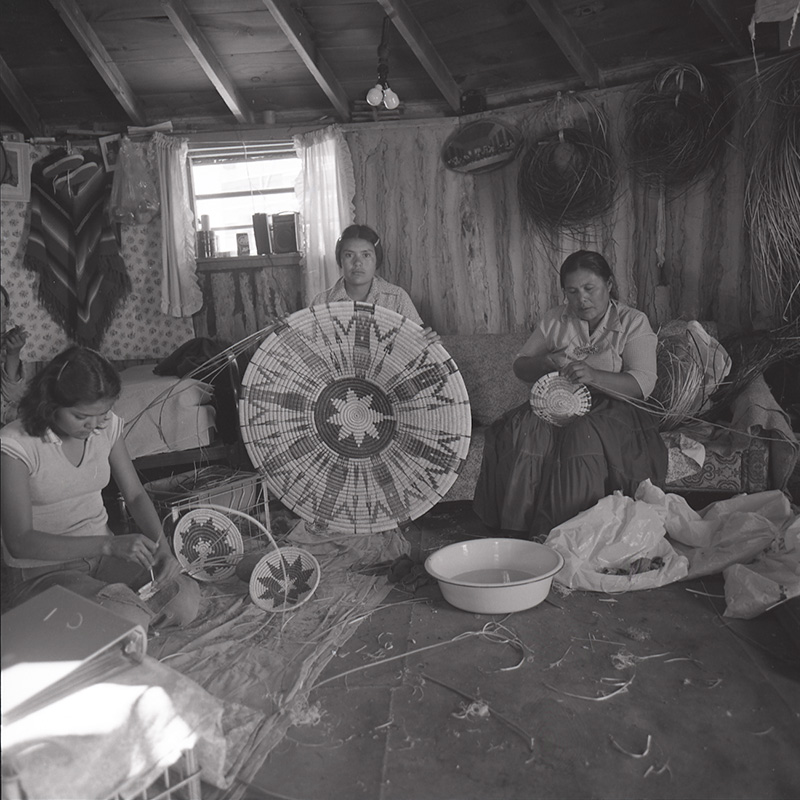 A black and white photo of a group of women in a hut making baskets.