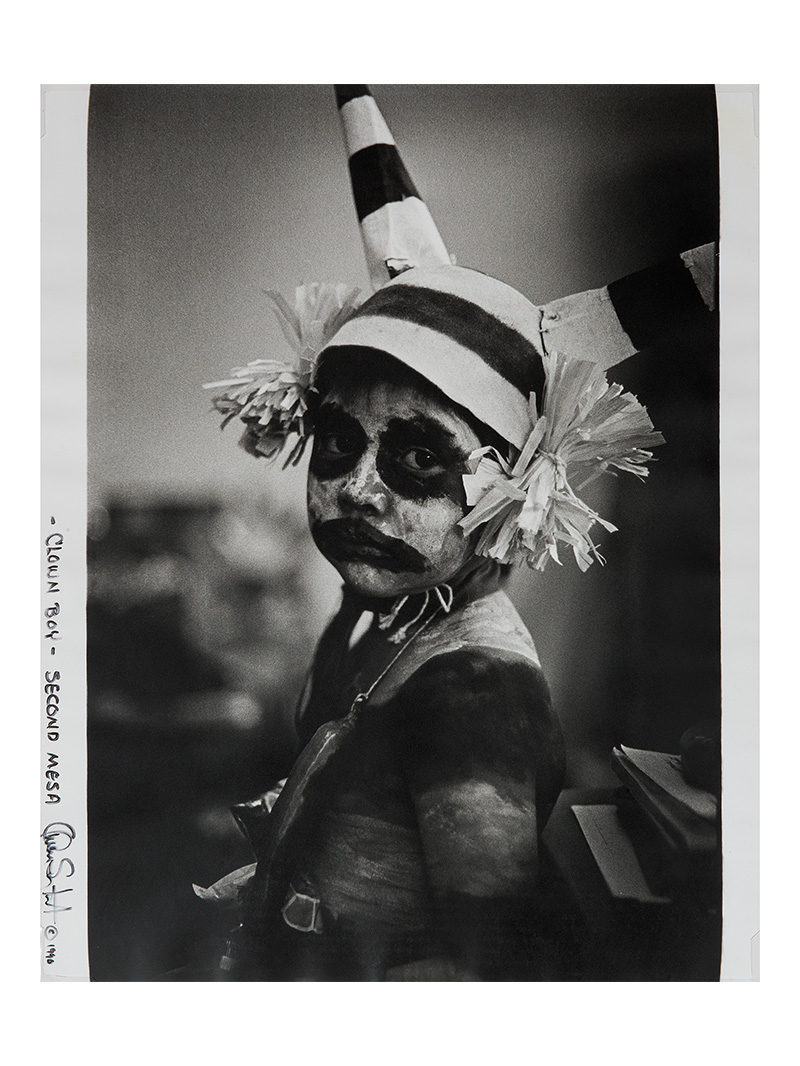 A black and white photograph of a child wearing a clown hat and makeup.