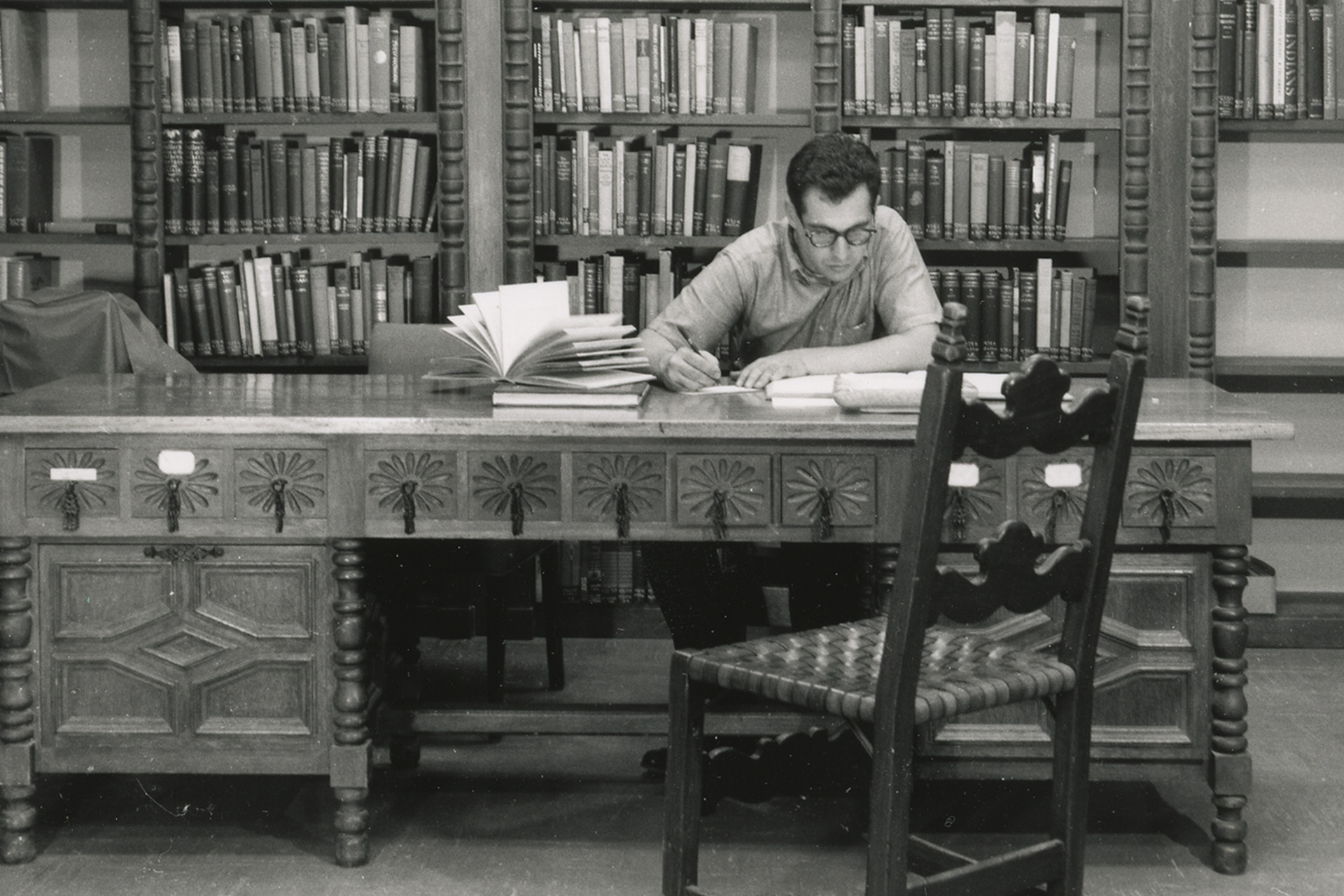 A black and white photo of a man wearing glasses sitting at a desk in a library.