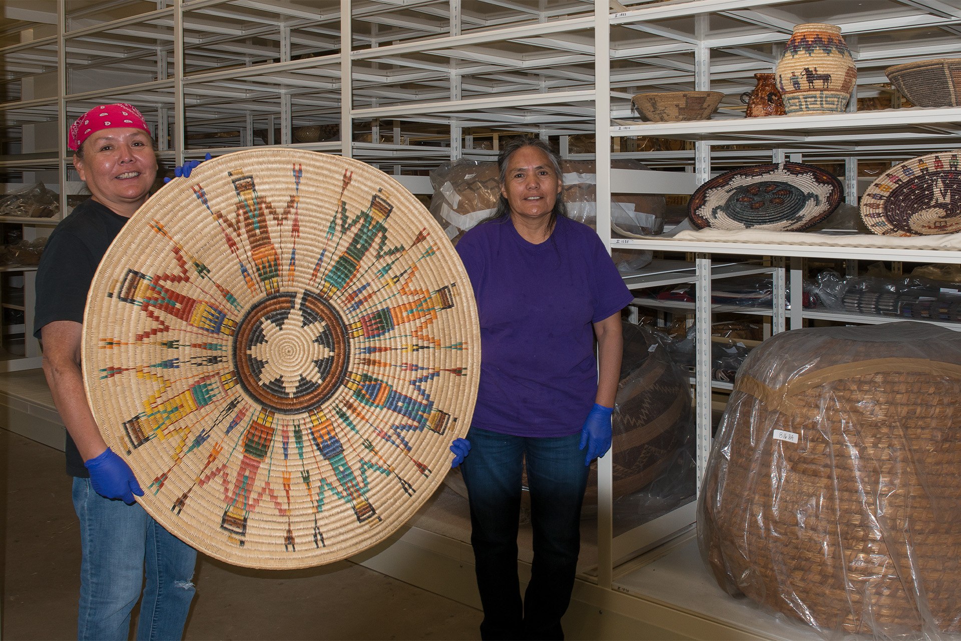 Two women hold an oversized Native pictorial basket in a storage room.