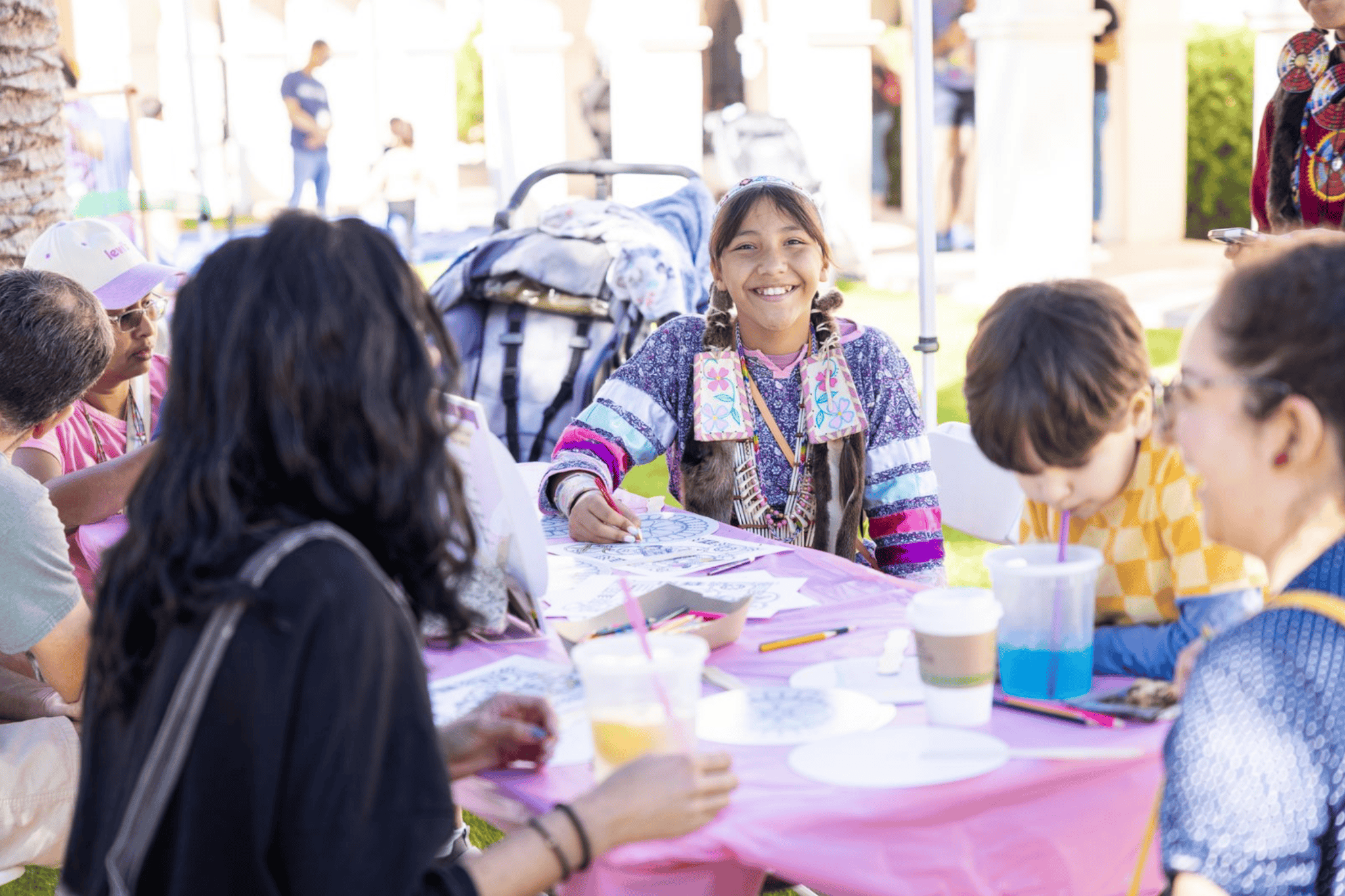 A group of people sits around a table outdoors, engaging in arts and crafts. A child in the center smiles at the camera, surrounded by colorful materials and beverages.