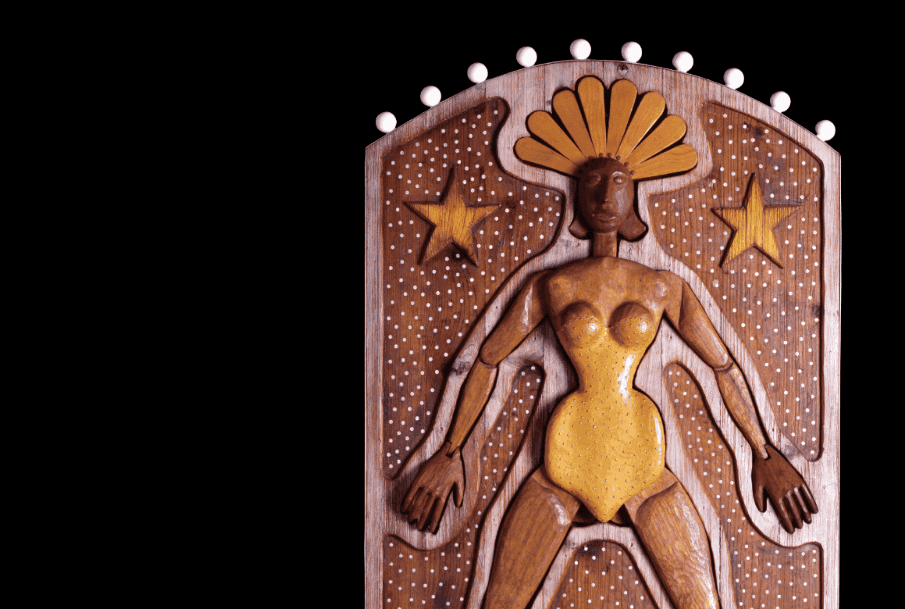 Wooden sculpture of a stylized female figure with a golden outfit, surrounded by stars and dots, against a black background.