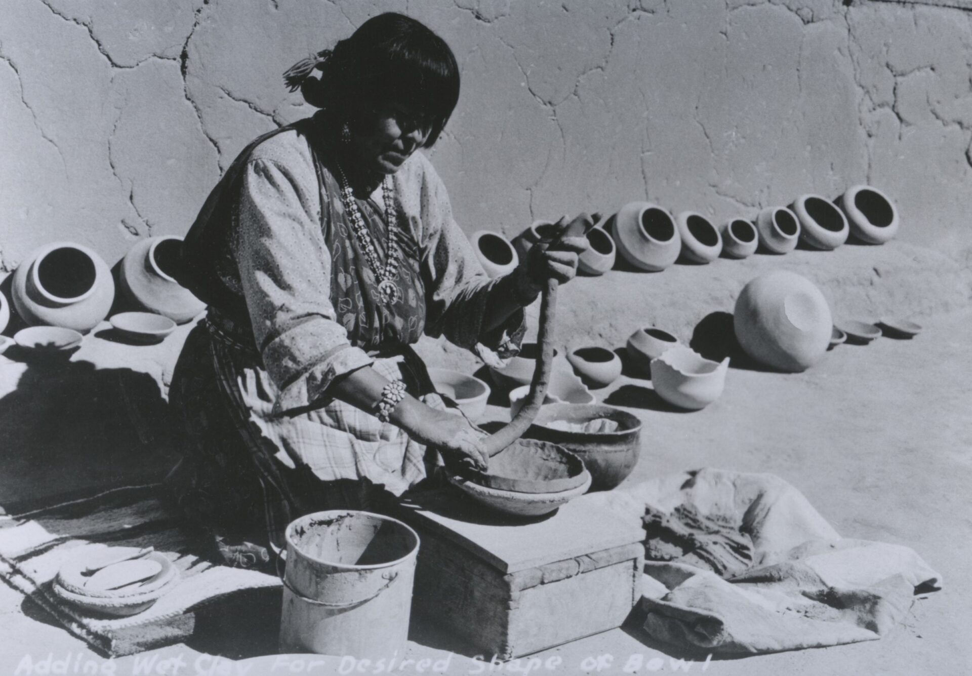 A woman shaping wet clay into a bowl with other pottery pieces lined up in the background.