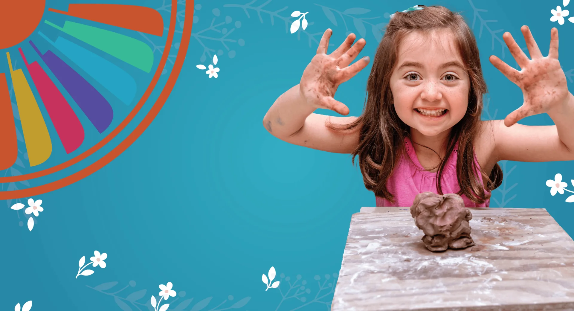 A cheerful young girl showing her paint-covered hands with a lump of brown clay on the table in front of her.