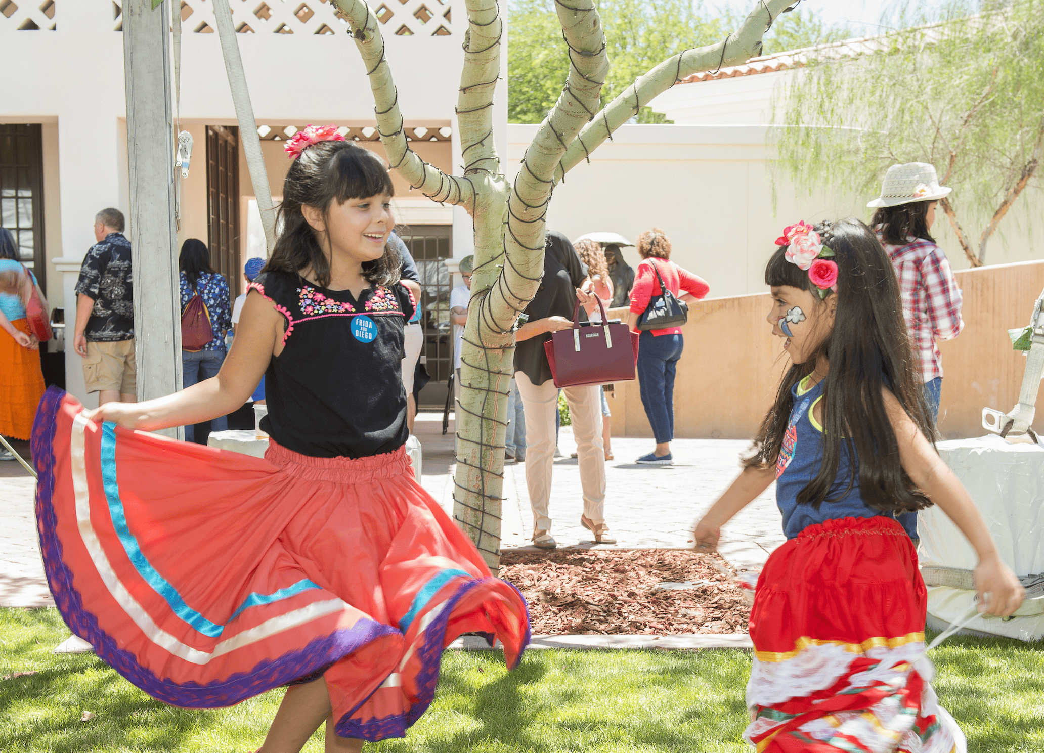 Two girls in colorful traditional dresses dancing under a tree at an outdoor event.