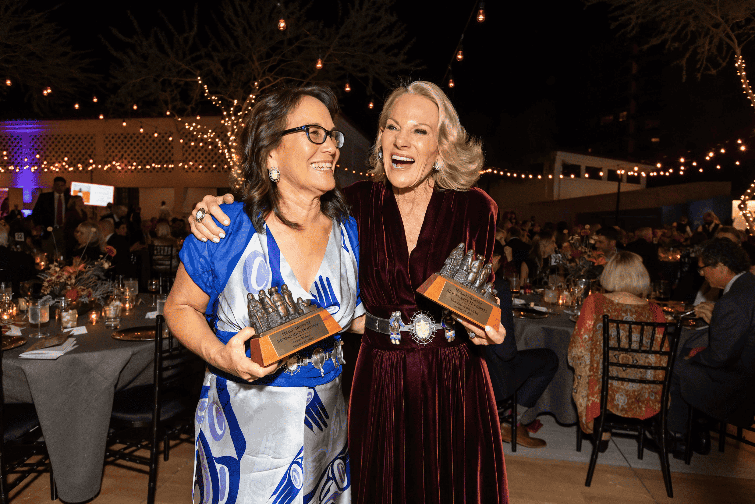 Two women smiling and holding awards at an evening moondance event with string lights in the background.