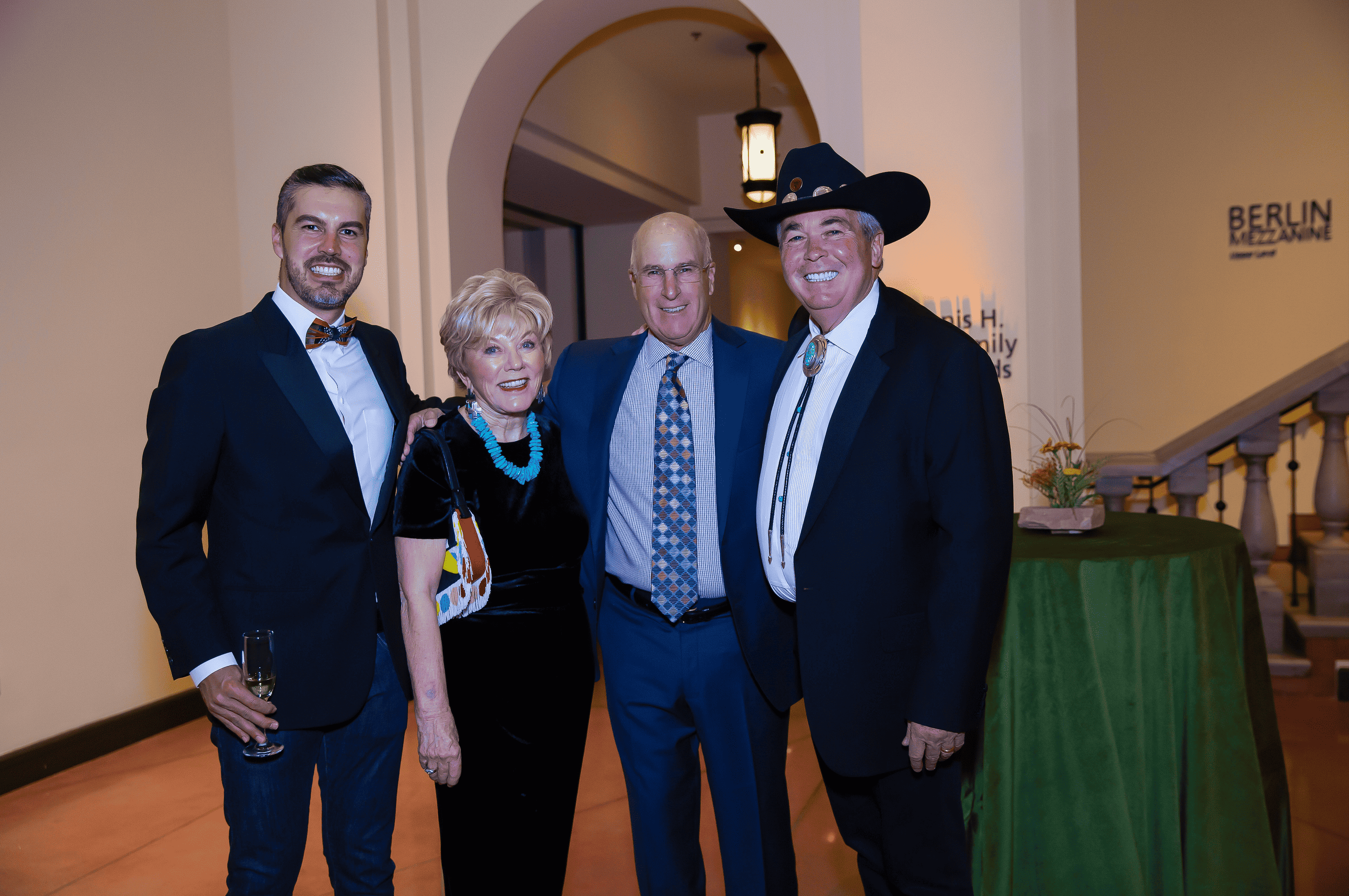 Four individuals at a moondance event, with two men wearing suits and one in a cowboy hat, standing with a woman adorned with turquoise jewelry.