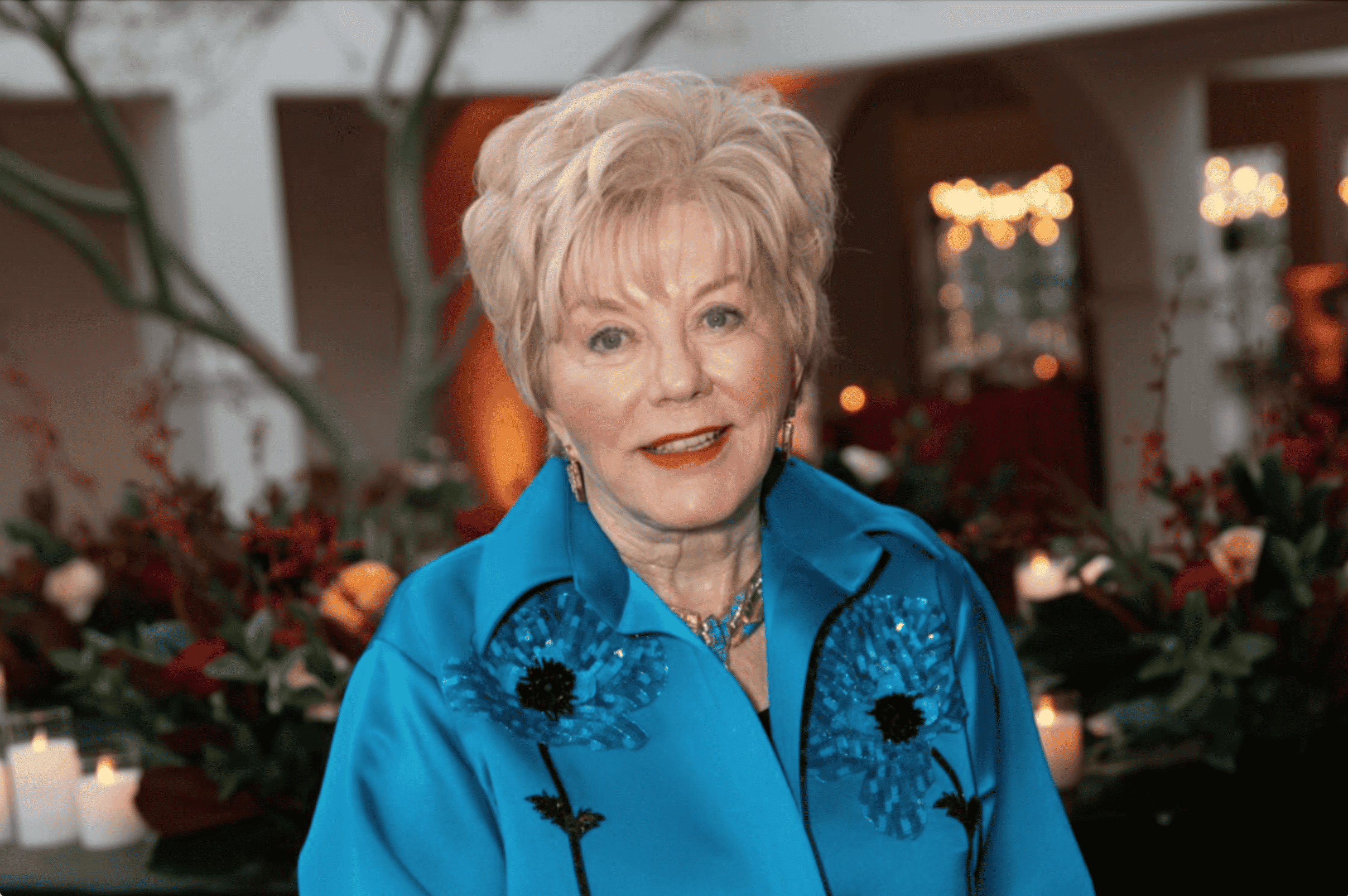 An elegant senior woman with styled blonde hair, wearing a bright blue blouse with moondance floral detailing, smiling at a formal event.