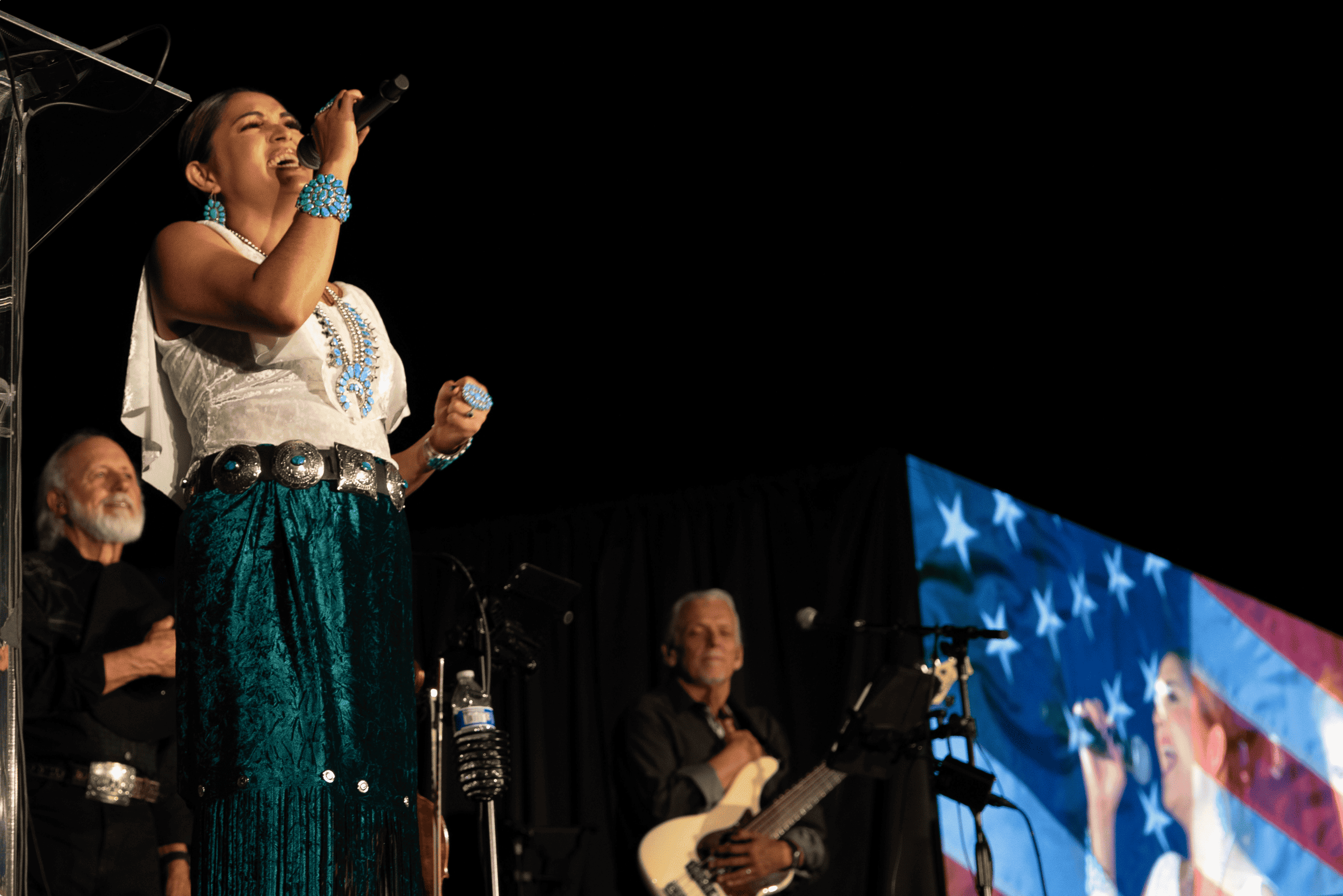 A female singer performing on stage with a microphone, accompanied by musicians, against the backdrop of a large screen displaying an American flag pattern during a moondance-themed set.