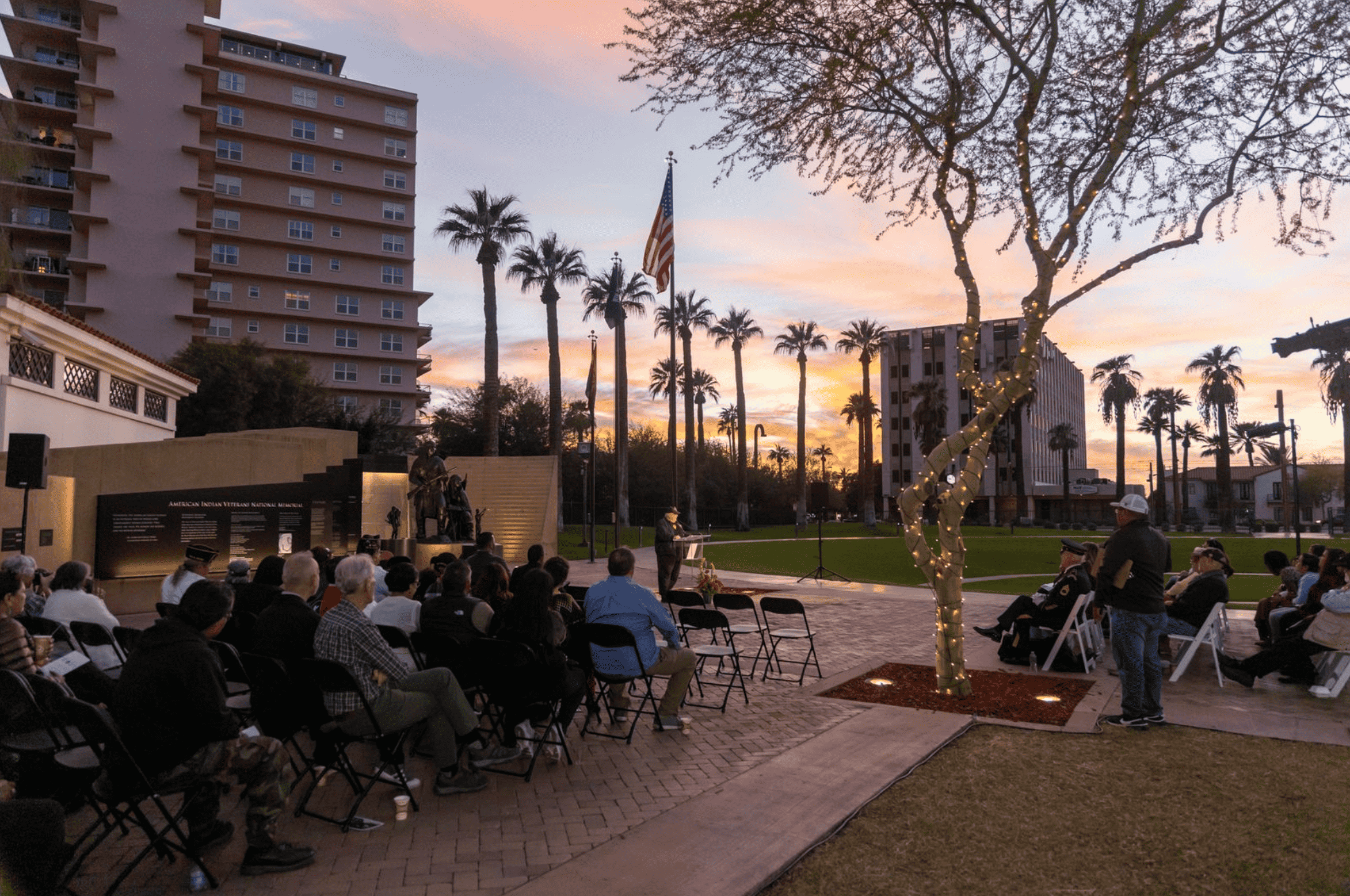 Outdoor event with attendees seated watching a speaker at dusk, surrounded by palm trees and a dramatic sunset, with city buildings in the background.