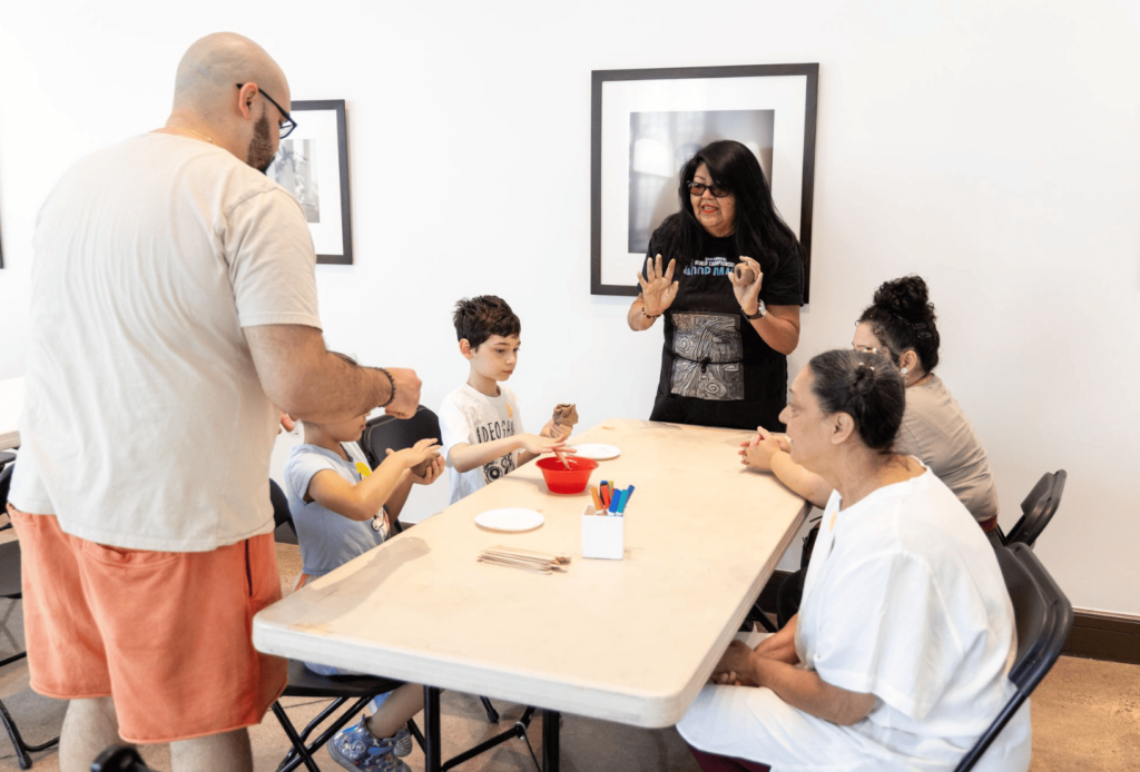 A family engages in a craft activity around a table while a woman stands and gestures as if explaining something.