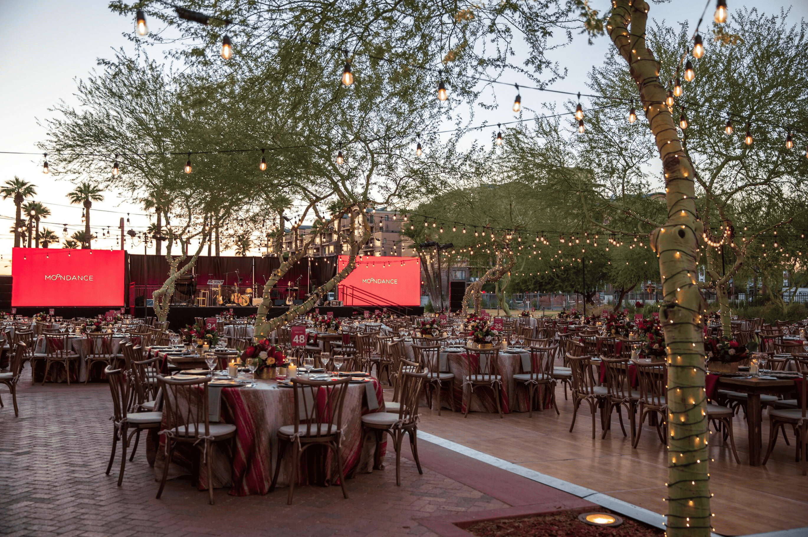Outdoor event setup with tables and string lights at dusk, featuring a stage with screens displaying 'phoenix' during a moondance.