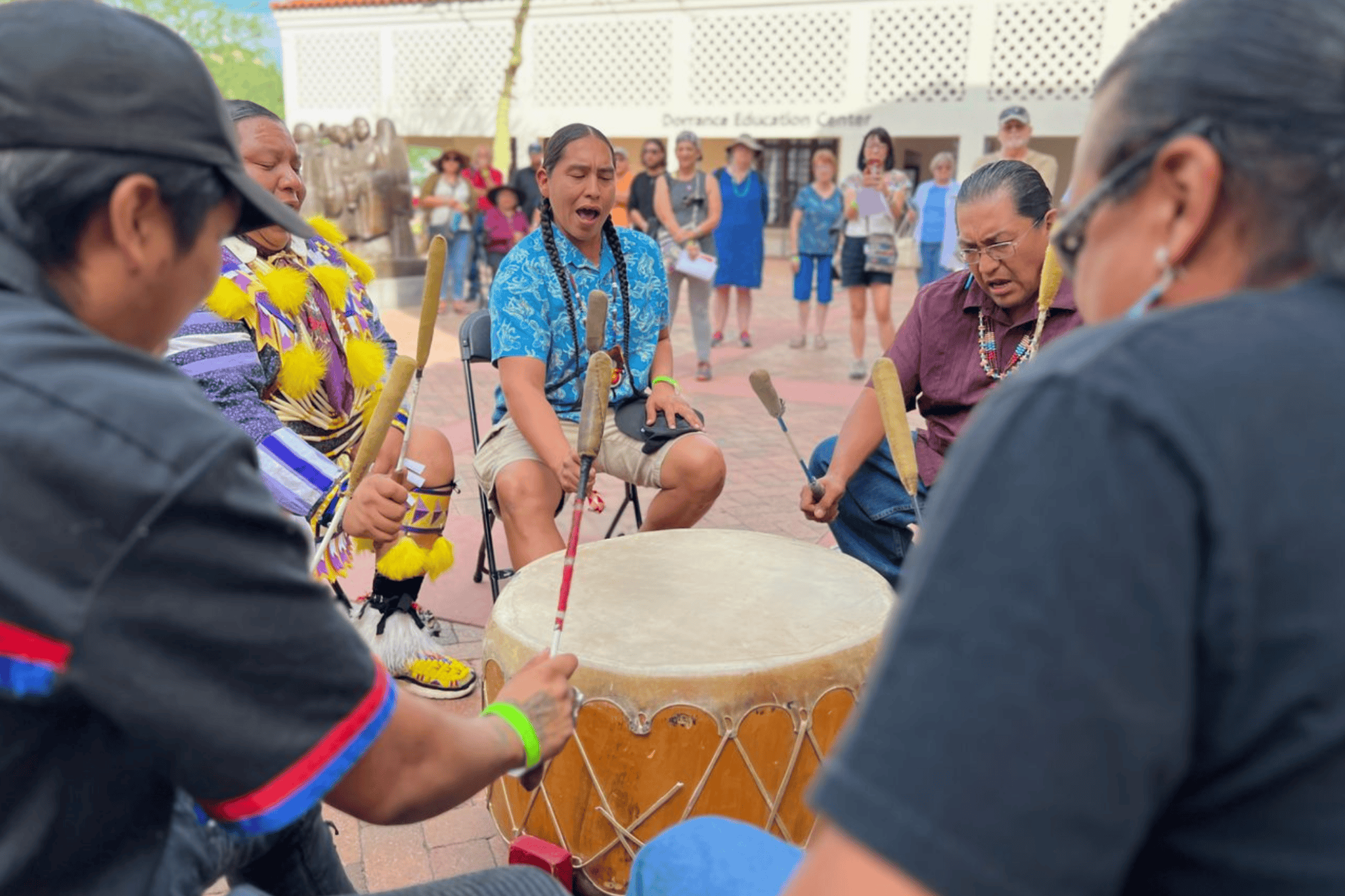 A group of people play traditional drums in a circle, with an audience watching in the background.