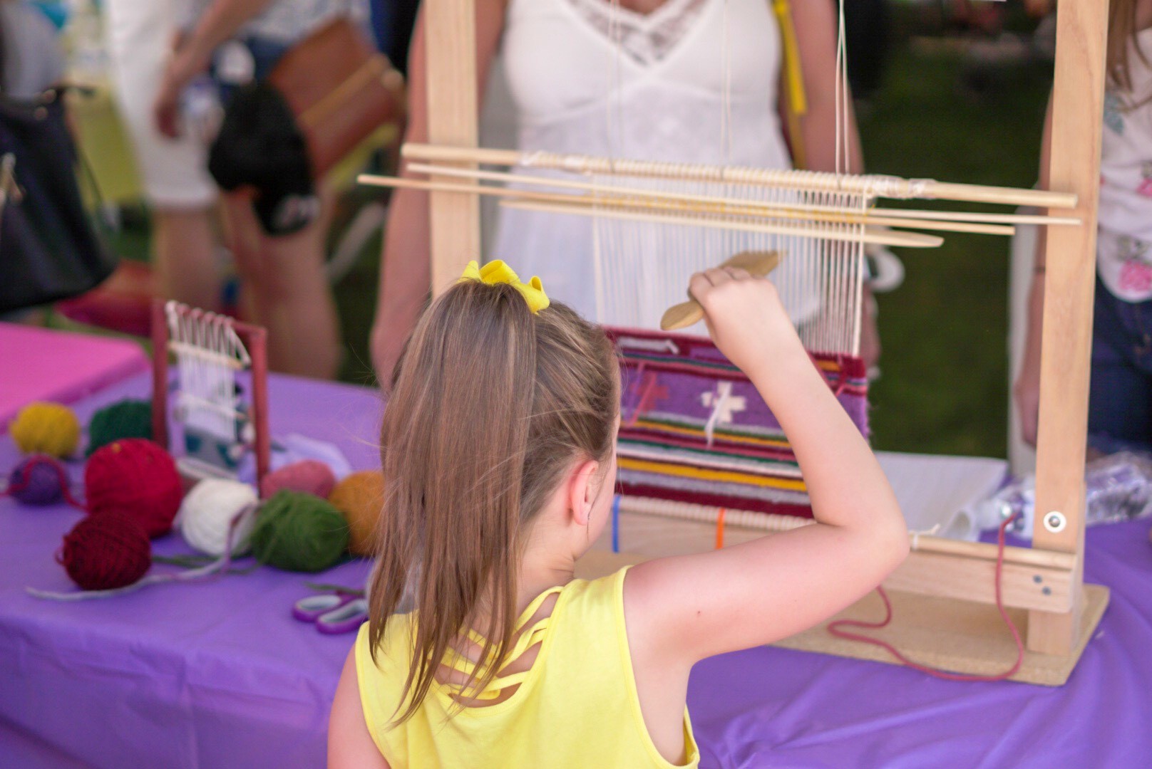 A young girl with a yellow hair bow weaves on a loom at a table with colorful yarn balls.