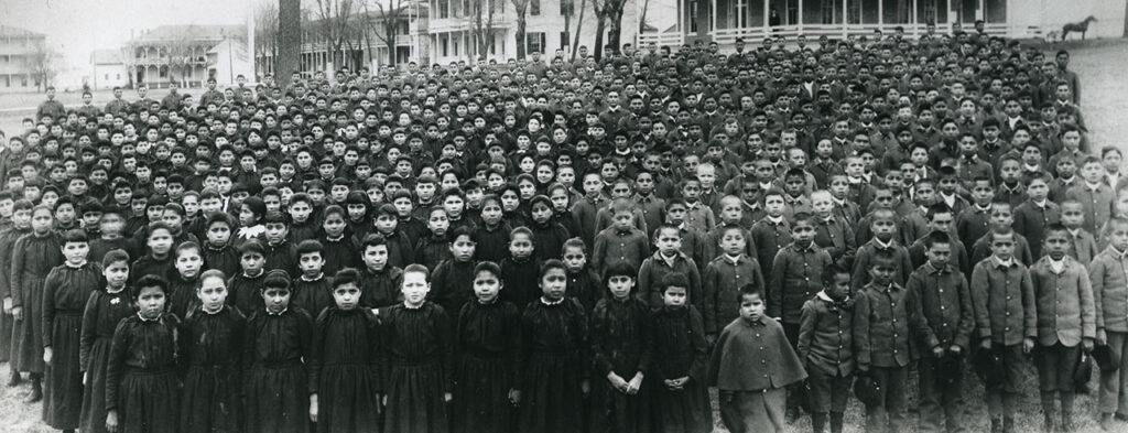 Black and white photo of school children in uniforms, black dresses on the girls and military style jackets and pants on the boys