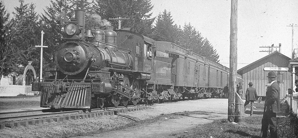 A steam locomotive pulls a passenger train past a small, rural station, with a few people standing nearby.