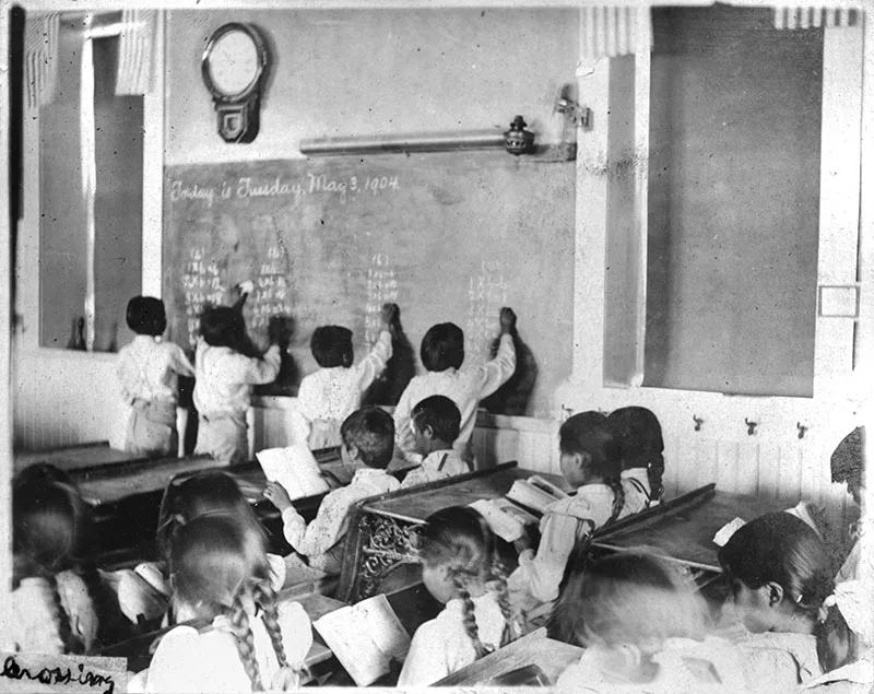 Students in a classroom circa 1904, some are seated at desks reading while a few stand at the blackboard working on assignments. A large clock and simple decor are visible on the walls.