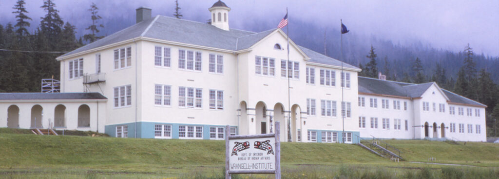 A large, two-story building with a sign in front reading "Mt. Edgecumbe High School Bureau of Indian Affairs." American and state flags are flying on poles in the foreground. Trees and mountains are in the background.