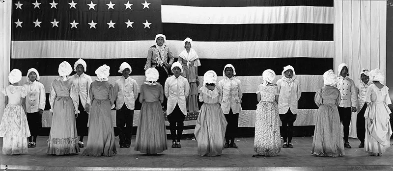 Group of people in colonial-era costumes standing in front of a large American flag with 13 stars. Some are facing forward, while others have their backs turned.