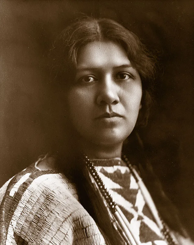 1900 black and white portrait of Angel de Cora, a young Native woman with bradied hear wearing a traditional beaded dress