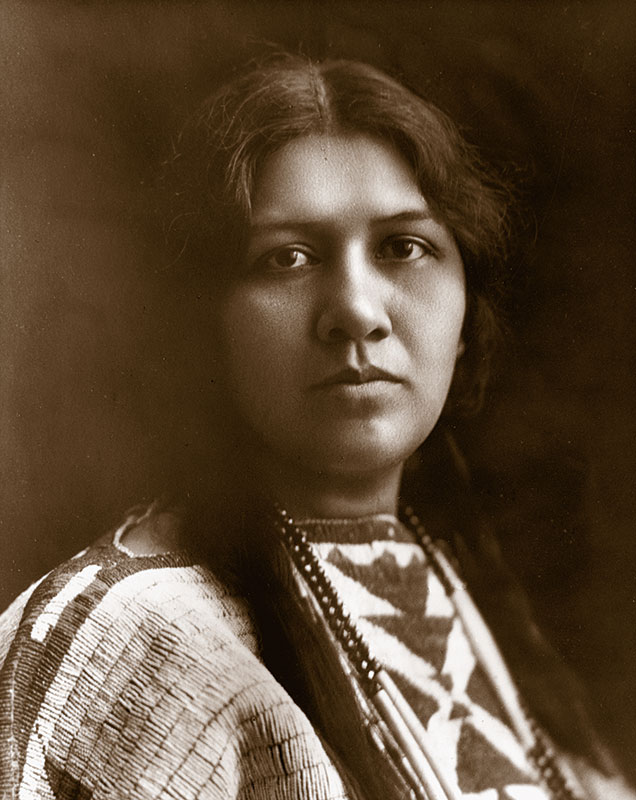 1900 black and white portrait of Angel de Cora, a young Native woman with bradied hear wearing a traditional beaded dress