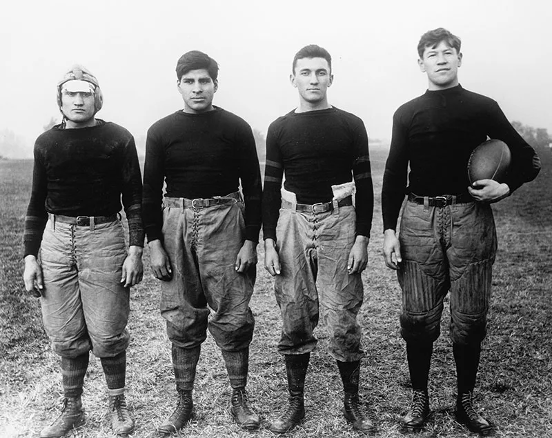 1912 black and white photo of 4 Native youth in early 20th C football uniforms posing for the camera. Jim Thorpe stands at far right holding a football.