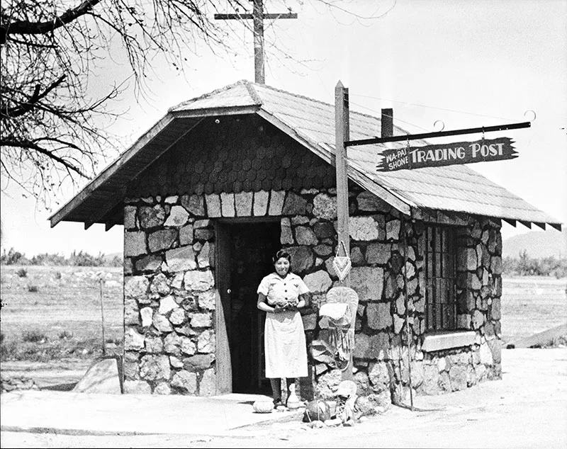A person stands in front of a small stone building labeled "Trading Post." There is a sign on the roof and various items displayed outside the entrance.