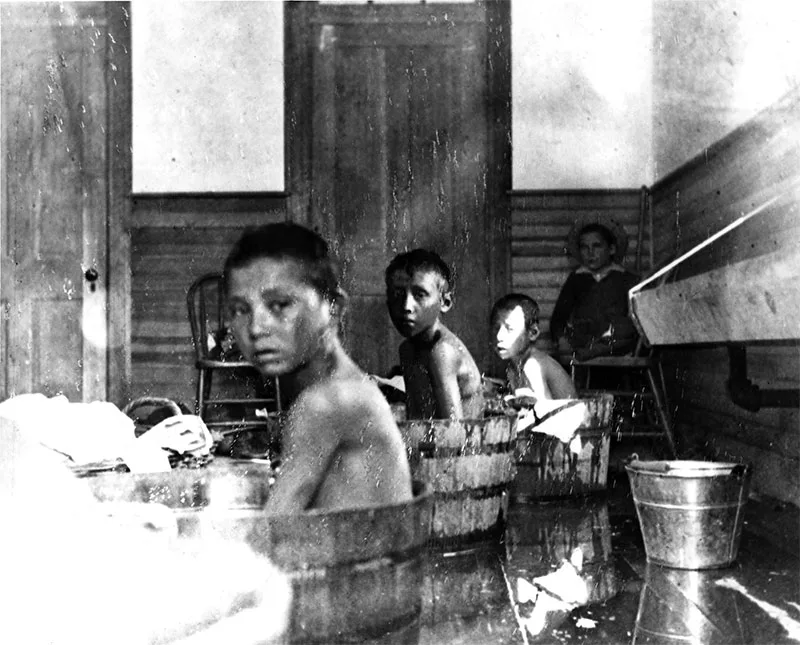 Children sit in wooden tubs filled with water, participating in a bath, while an adult supervises in a room with wooden walls and doors.