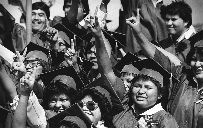 1984 candid black and white photo of Native youth in graduation caps and gowns smiling and holding their right arms in the air with their pointer fingers raised.
