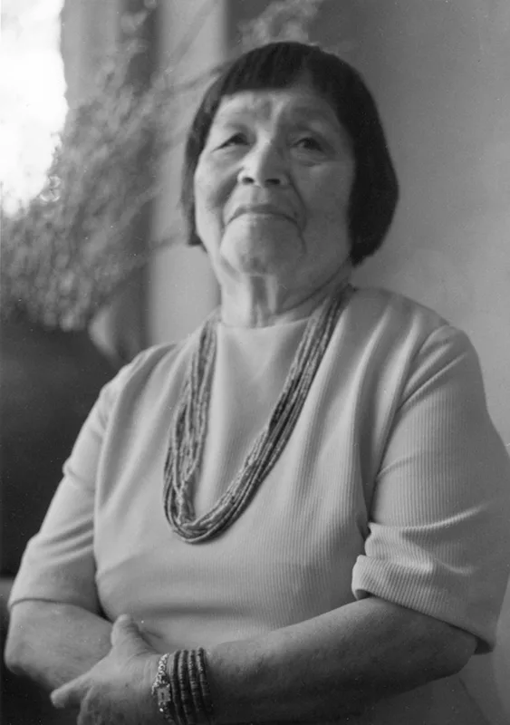 Black and white photo of an elderly woman with short hair, wearing a light-colored dress and multiple necklaces, looking slightly upwards with a contemplative expression.