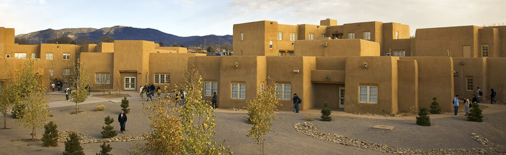A wide view of an adobe-style building complex with multiple people walking on the paths around it. Small trees and shrubs are in the surrounding landscape, and mountains can be seen in the background.