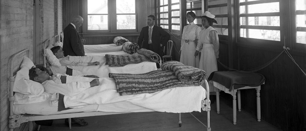 Black and white photo of a turn of the century infirmary. Two doctors and 2 nurses stand by beds with young men.
