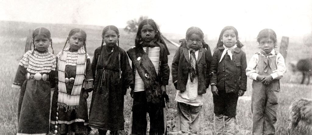 A black and white photo of seven young children standing in a line outdoors, labeled "Sioux 1891." They are wearing traditional clothing and stand in a field with blurred background elements.