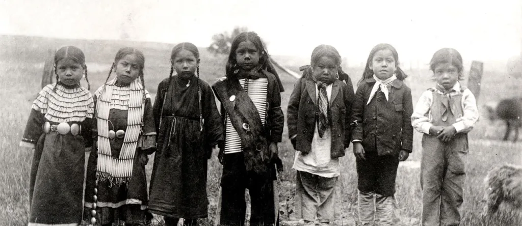 A black and white photo of seven young children standing in a line outdoors, labeled "Sioux 1891." They are wearing traditional clothing and stand in a field with blurred background elements.
