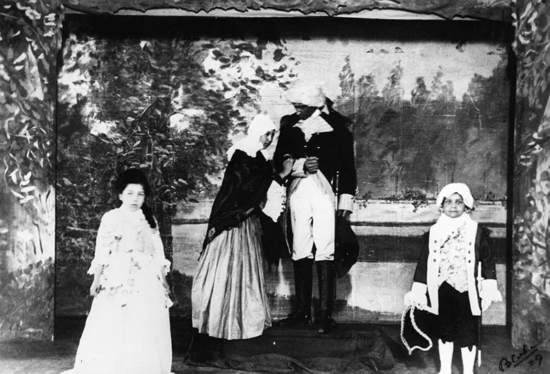 Three actors in historical costumes perform a stage scene with painted backdrop; two are conversing while the third looks on. Another costumed actor stands to the side holding props.