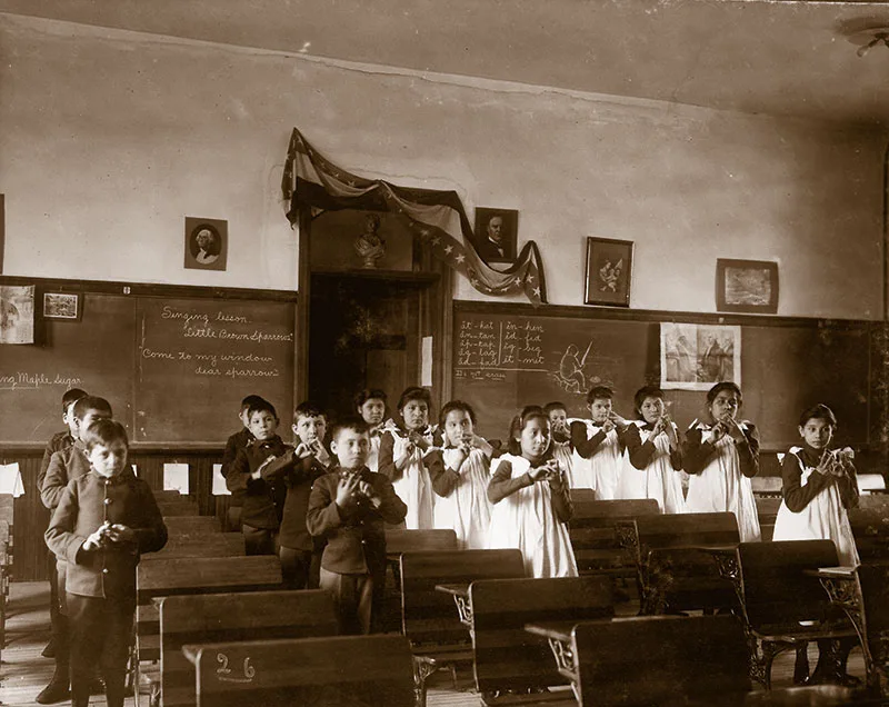 A group of children stands playing recorders in a classroom, wearing formal attire. The classroom has chalkboards with writing and pictures hanging on the walls.