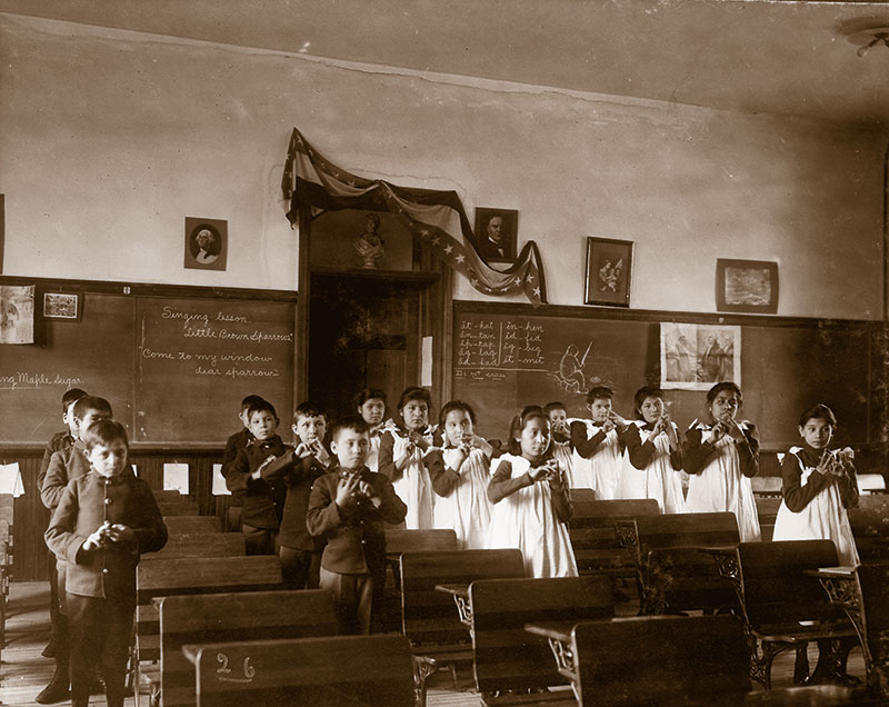 A group of children stands playing recorders in a classroom, wearing formal attire. The classroom has chalkboards with writing and pictures hanging on the walls.