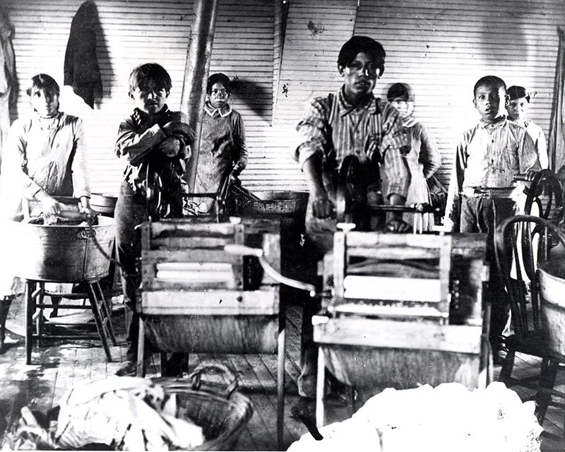 1901 black and white photo of young Native boys in a laundry room in front of wringing machines and wash tubs
