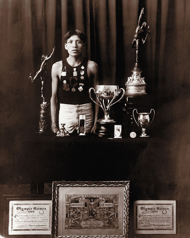 1908 black and white photo of young Native male in track and field shorts and sleeveless top posing with trophies and medals and framed awards including 2 1908 Olympics certificates.