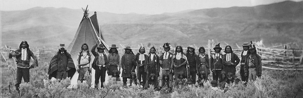 Black and white photograph of a group of Native American men and children standing in front of a tipi and a wooden structure, set against a mountainous landscape.