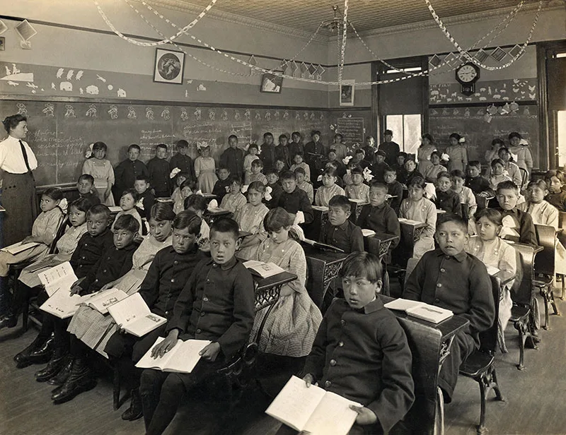 Historical classroom scene with a large group of children seated at desks, holding open books, and a teacher standing on the left. Many students are looking towards the camera. Chandeliers and chalkboards are visible.