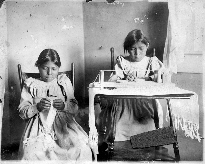 1905 black and white photo of 2 young Native girls working on beading. One girl sits at a desk with a traditional bead loom and the other sits beside with indistinct fabric in her hand.