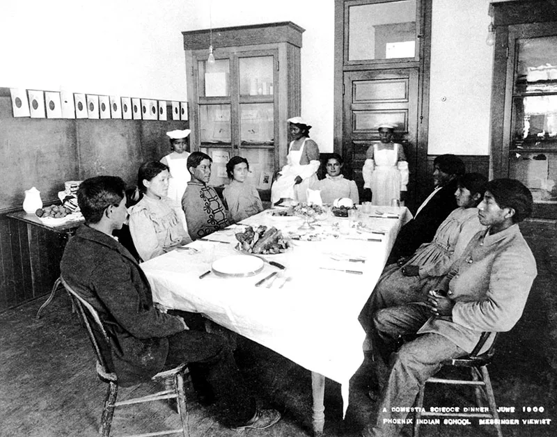 1900 black and white photo of Native teenagers sitting formally at a dinner table with 3 females in domestic service uniforms behind.